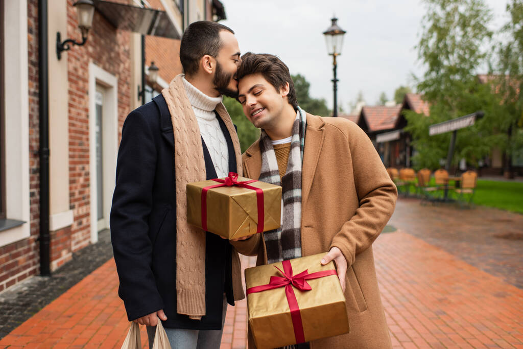 γενειοφόρος γκέι άντρας φιλάει το κεφάλι του ευτυχισμένου αγοριού κρατώντας χριστουγεννιάτικα δώρα στο δρόμο της πόλης - Φωτογραφία, εικόνα