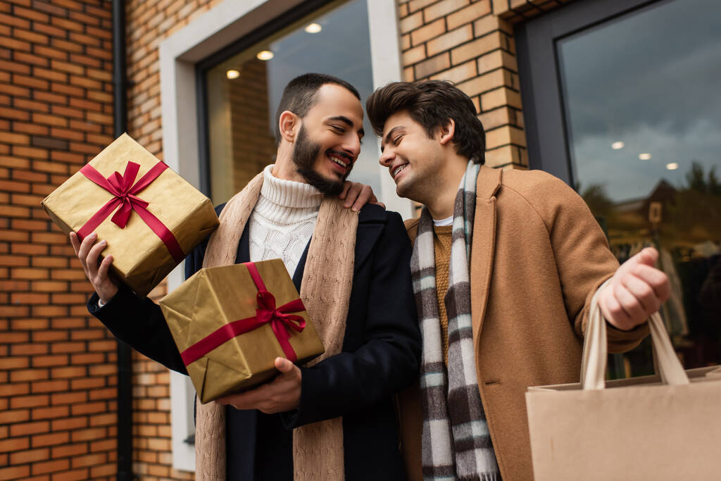 μοντέρνο γκέι ζευγάρι με χριστουγεννιάτικα δώρα και τσάντα ψώνια χαμογελώντας κοντά κατάστημα με προθήκες στο δρόμο της πόλης - Φωτογραφία, εικόνα