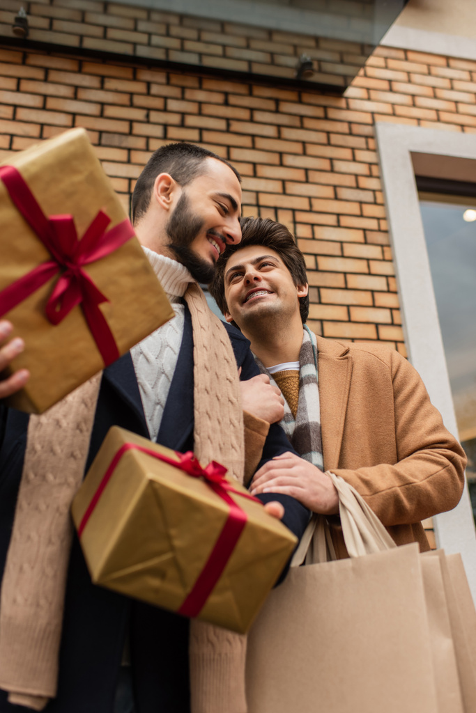 niski kąt widzenia wesoły gej mężczyźni z prezenty świąteczne i torby na zakupy w pobliżu budynku na ulicy - Zdjęcie, obraz