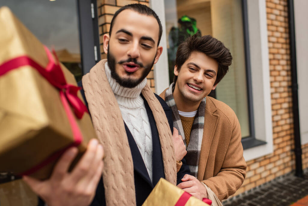 Şaşkın gay elinde bulanık bir Noel hediyesi kutusu tutuyor neşeli erkek arkadaşının yanında ve şehir sokağındaki dükkanda - Fotoğraf, Görsel