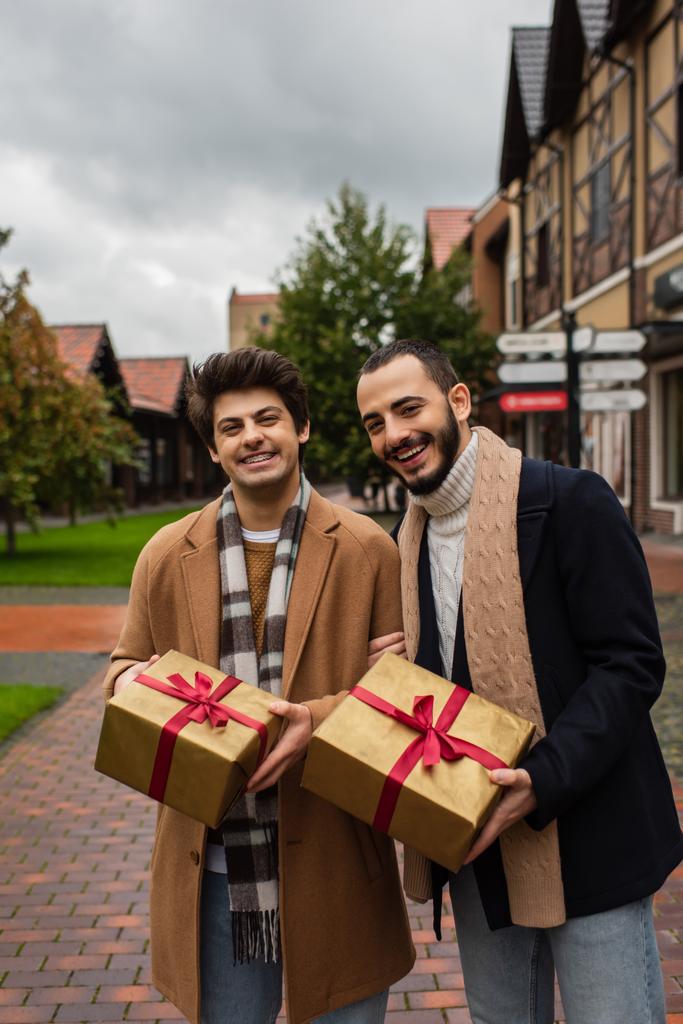 excited and fashionable gay couple with Christmas gift boxes looking at camera on city street - Photo, Image