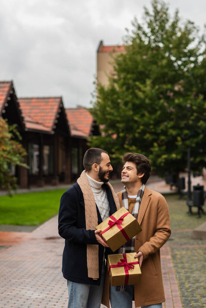 trendy gay paar met kerstcadeaus glimlachen naar elkaar op straat - Foto, afbeelding