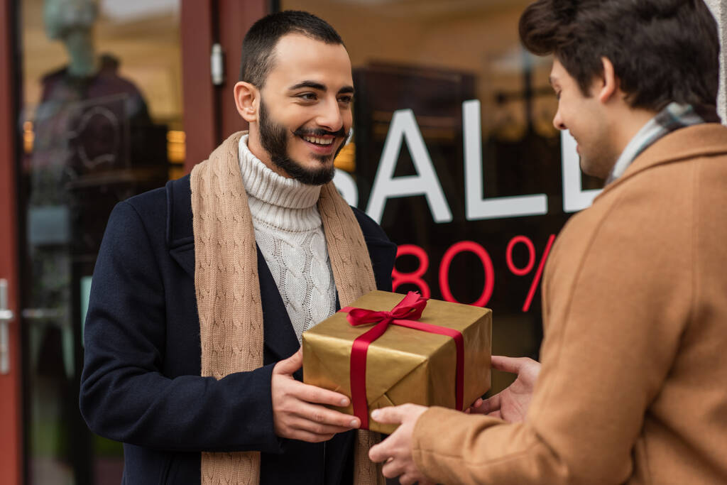 heureux barbu homme tenant cadeau de Noël près flou copain et vitrine avec vente lettrage  - Photo, image