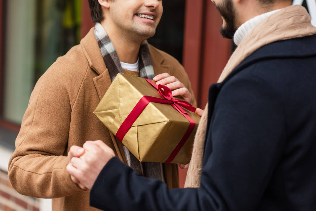 vista parcial de la sonriente pareja gay cogida de la mano cerca de la caja de regalo en la calle borrosa - Foto, Imagen