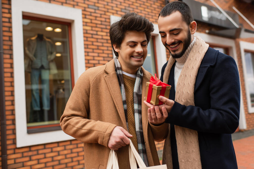 joven gay hombre con compras bolsa celebración regalo caja cerca feliz barbudo novio y tienda en borrosa fondo - Foto, Imagen