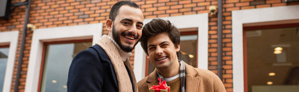 onnellinen homo miehet tilalla pieni lahjapaketti lähellä ostoksia hämärtynyt tausta, banneri - Valokuva, kuva