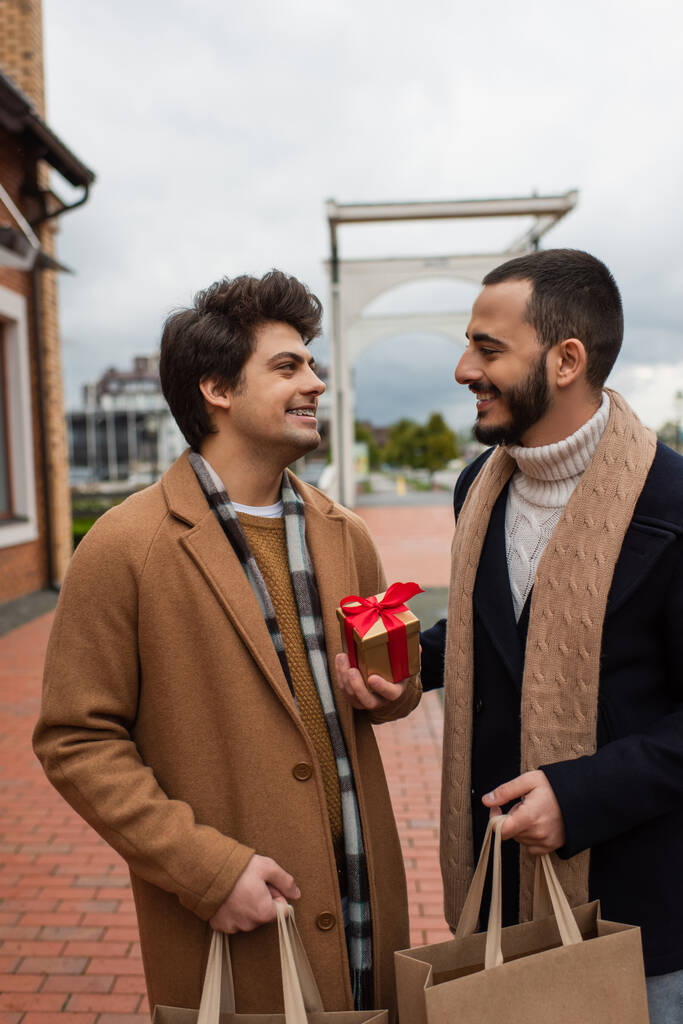 mladý gay muž s nákupními taškami a vánoční dárek při pohledu na šťastný vousatý přítel  - Fotografie, Obrázek