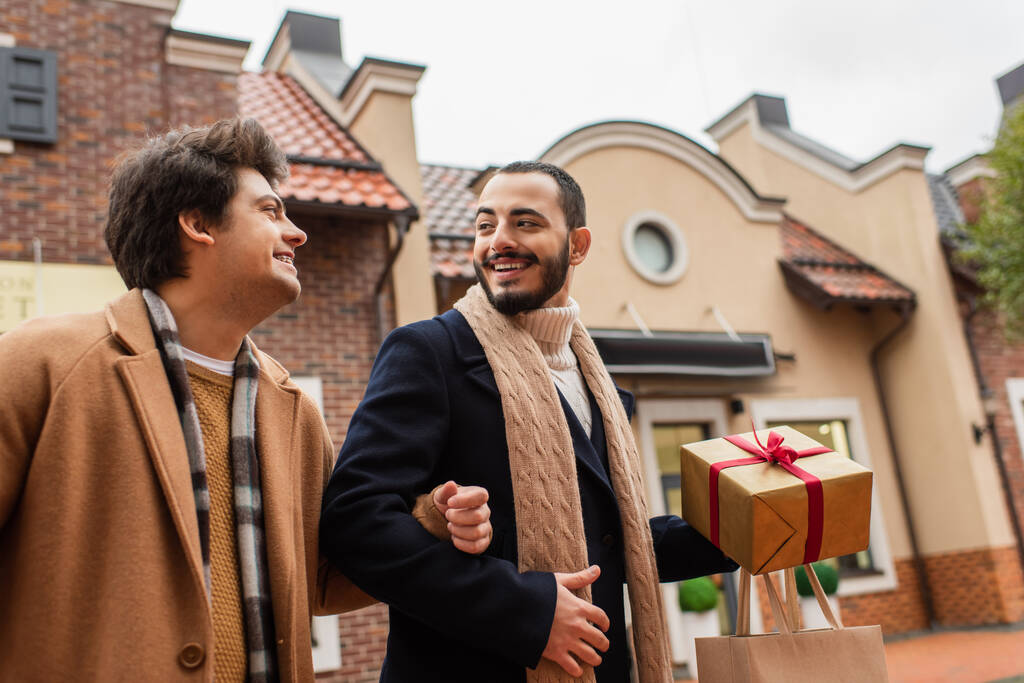 bearded gay man with shopping bag and Christmas present looking at young boyfriend on urban street - Photo, Image