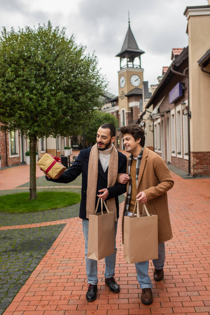 full length of trendy gay partners with shopping bags looking at gift box on urban street - Photo, Image