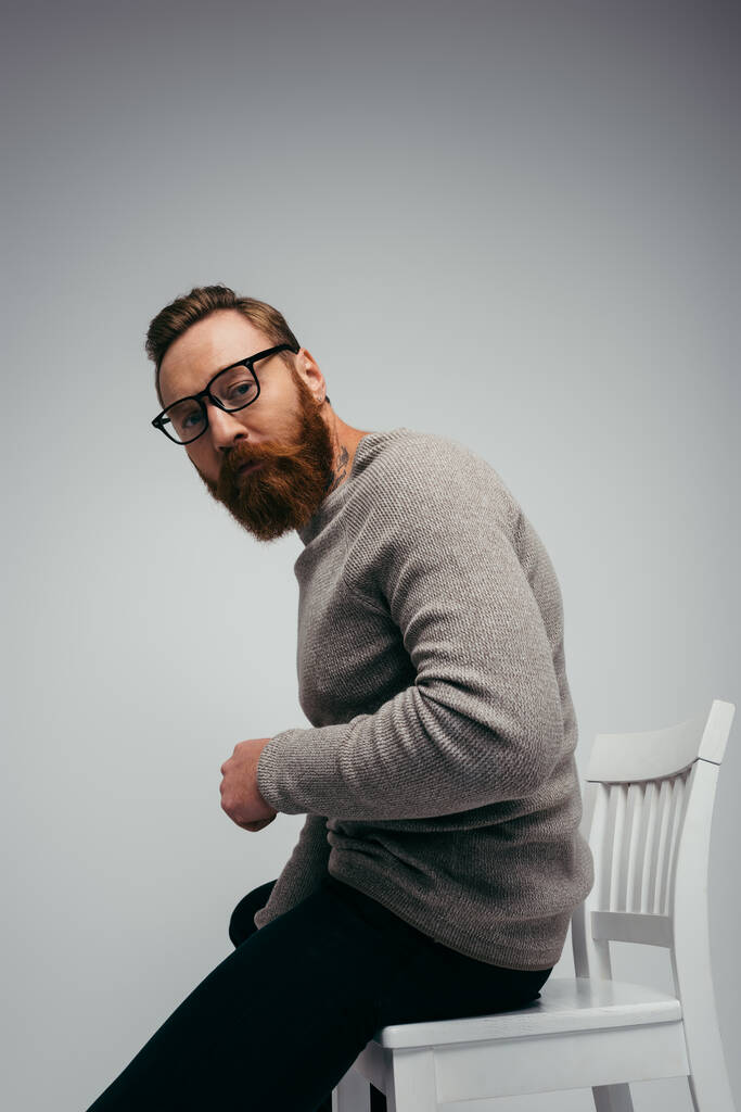 Стильний бородатий чоловік в окулярах сидить на білому стільці ізольовано на сірому
  - Фото, зображення
