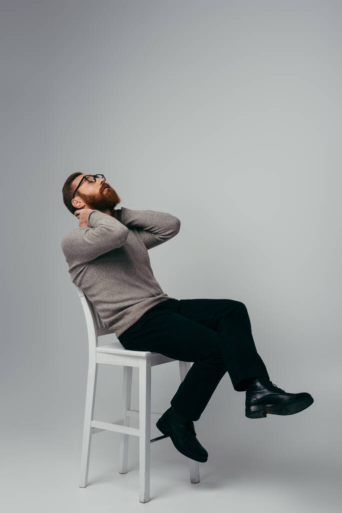 Comprimento total do modelo em suéter e óculos tocando no pescoço enquanto sentado na cadeira em fundo cinza  - Foto, Imagem