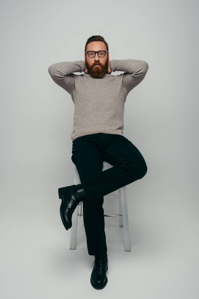 Longitud completa de hombre con estilo en gafas que tocan el cuello y mirando a la cámara mientras está sentado en la silla sobre fondo gris  - Foto, imagen