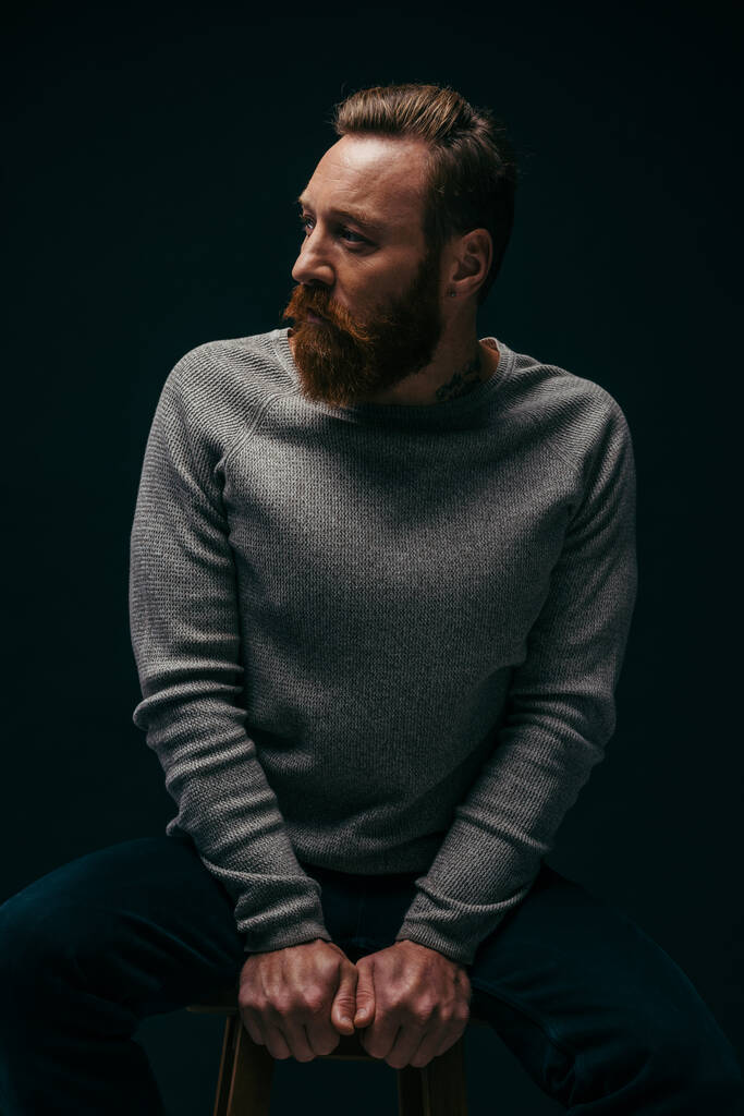 Hombre de moda en suéter mirando hacia otro lado mientras está sentado en la silla aislado en negro con sombra  - Foto, Imagen
