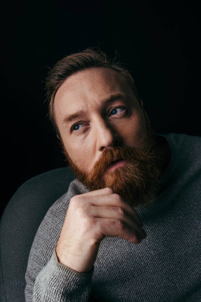 黒に隔離されたアームチェアに座っているセーターの髭の男の肖像  - 写真・画像