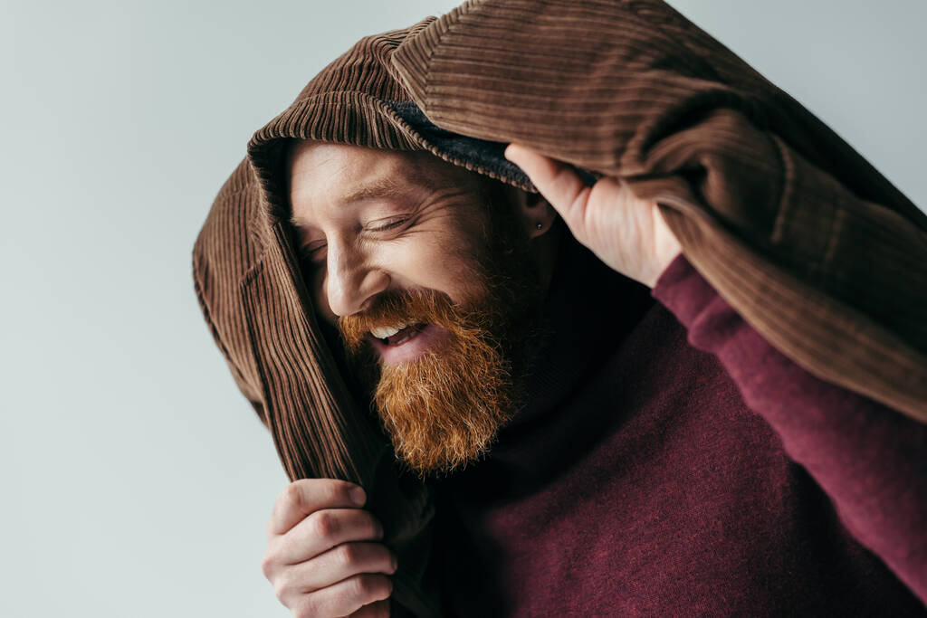 счастливый мужчина с закрытыми глазами и коричневым блейзером на голове, улыбающийся изолированно на сером  - Фото, изображение