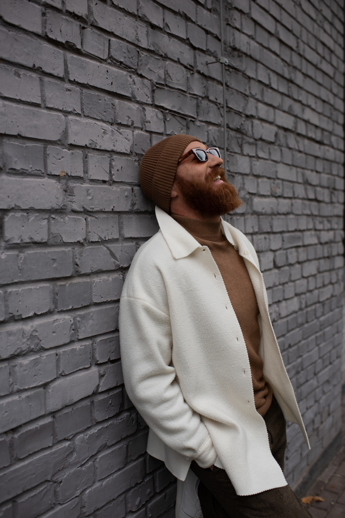 Tuğla duvara dayanmış bereli ve güneş gözlüklü neşeli ve sakallı bir adam.  - Fotoğraf, Görsel