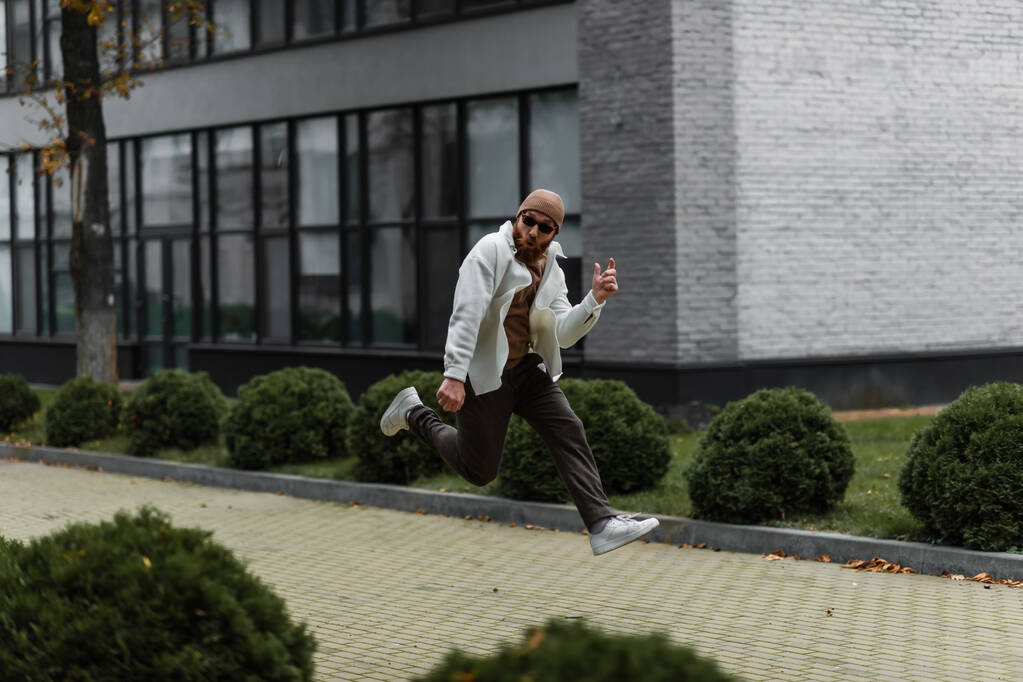 volledige lengte van de bebaarde man in muts en trendy zonnebril springen in de buurt van modern gebouw  - Foto, afbeelding