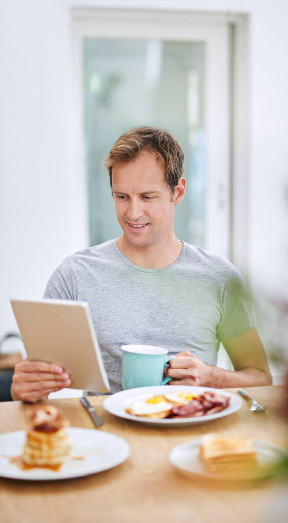Lee sus noticias en línea. un hombre guapo usando su tableta mientras desayuna en casa - Foto, imagen
