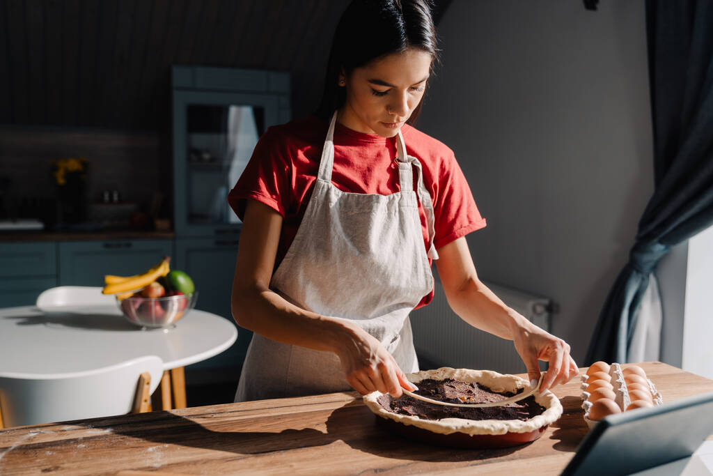 Jeune femme hispanique portant tablier faisant tarte dans la cuisine à la maison - Photo, image
