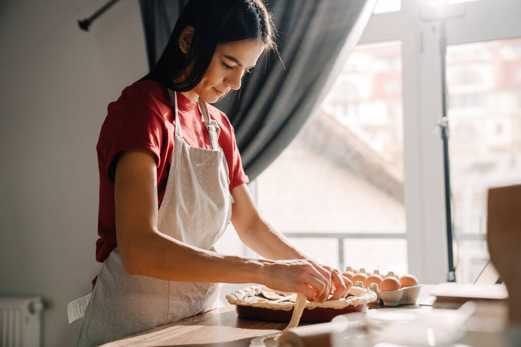 Νεαρή Ισπανίδα μελαχρινή γυναίκα φοράει ποδιά μαγειρεύοντας και φτιάχνοντας πίτα στην κουζίνα στο σπίτι - Φωτογραφία, εικόνα