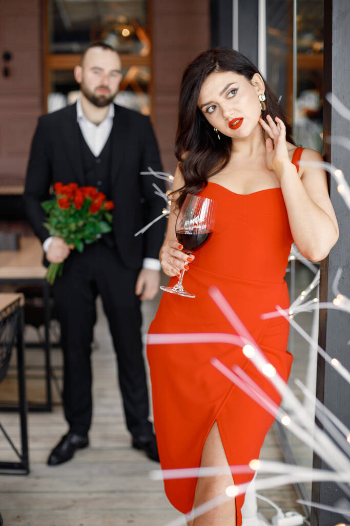 Koncentrálj a vonzó, piros ruhás fiatal nőre egy romantikus vacsorára. Egy férfi áll mögötte egy csokor vörös rózsával. Gyönyörű szerető pár együtt tölti az idejét a modern étteremben.. - Fotó, kép