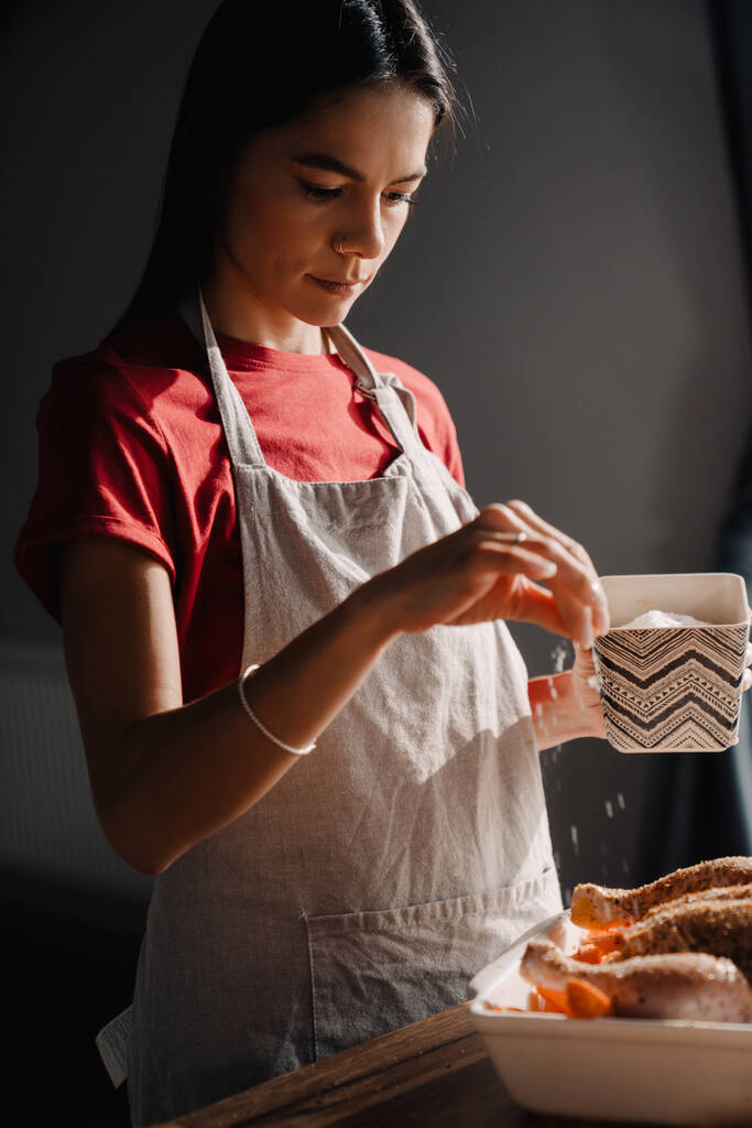 Mujer hispana joven usando delantal cocinando pollo en la cocina en casa - Foto, Imagen