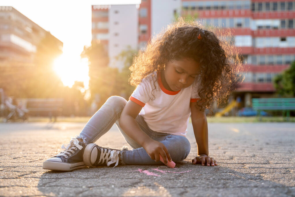 Συμπυκνωμένο κοριτσάκι που κάθεται στο πλακόστρωτο έδαφος και ζωγραφίζει με μια χρωματιστή κιμωλία - Φωτογραφία, εικόνα