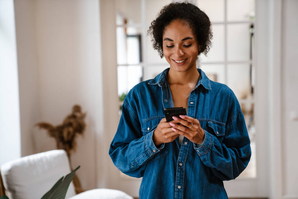 Giovane donna nera che indossa camicia di jeans sorridente e utilizzando il cellulare a casa - Foto, immagini