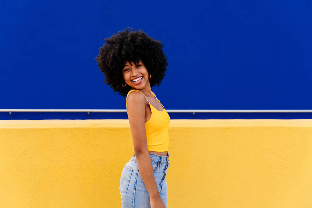 Krásná mladá šťastná africká žena s afro kudrnatý účes procházky ve městě - Veselé černošky student portrét na barevné pozadí zdi - Fotografie, Obrázek