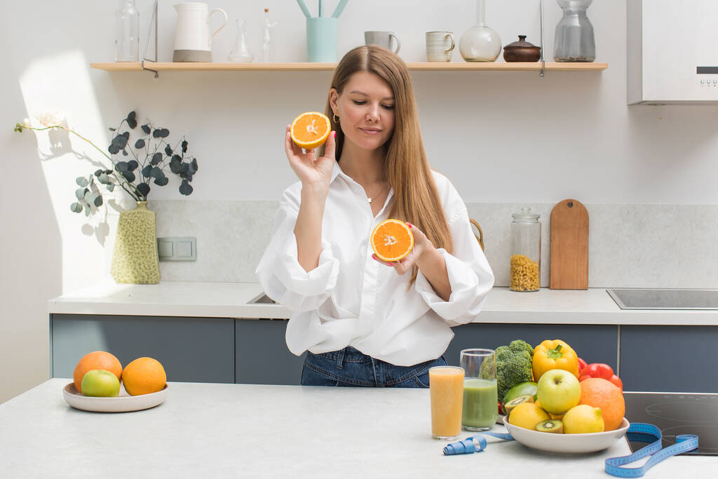 A young blond woman holds orange halves in her hands in the kitchen next to a table with fruits. The concept of diet, healthy eating. - Photo, Image