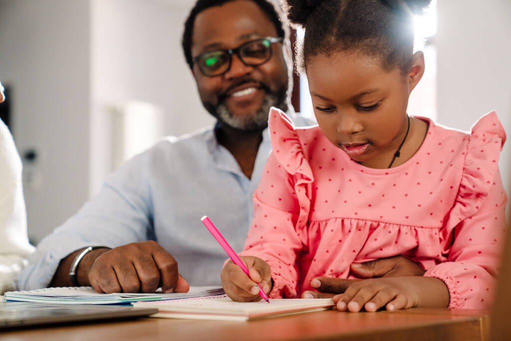Αφρικανική αμερικανική κοριτσάκι σχέδιο σε σημειωματάριο με δείκτη, ενώ κάθεται με τους γονείς από το τραπέζι στο σπίτι - Φωτογραφία, εικόνα