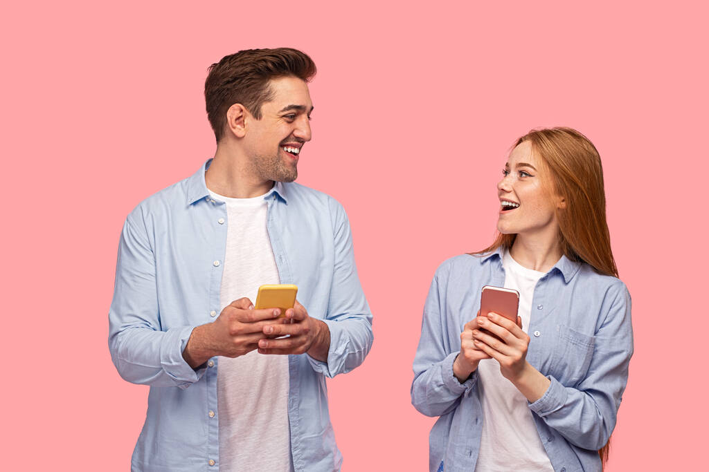 Fröhliche junge Freundin und Freund in ähnlichen Kleidern benutzen Handys und schauen einander lächelnd vor rosa Hintergrund an - Foto, Bild