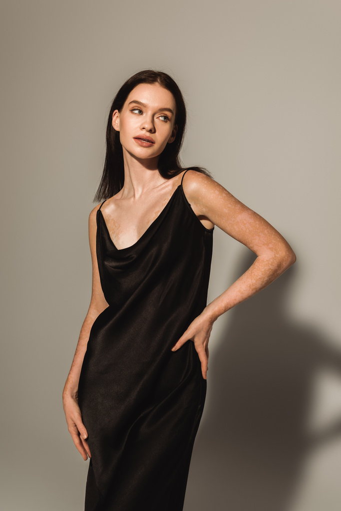 Κομψό μοντέλο με λεύκη σε μαύρο μεταξωτό φόρεμα στέκεται με το χέρι στο ισχίο σε γκρι φόντο  - Φωτογραφία, εικόνα