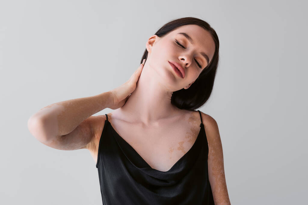 Νεαρή γυναίκα με λεύκη αγγίζοντας το λαιμό και κλείνοντας τα μάτια απομονώνονται σε γκρι  - Φωτογραφία, εικόνα