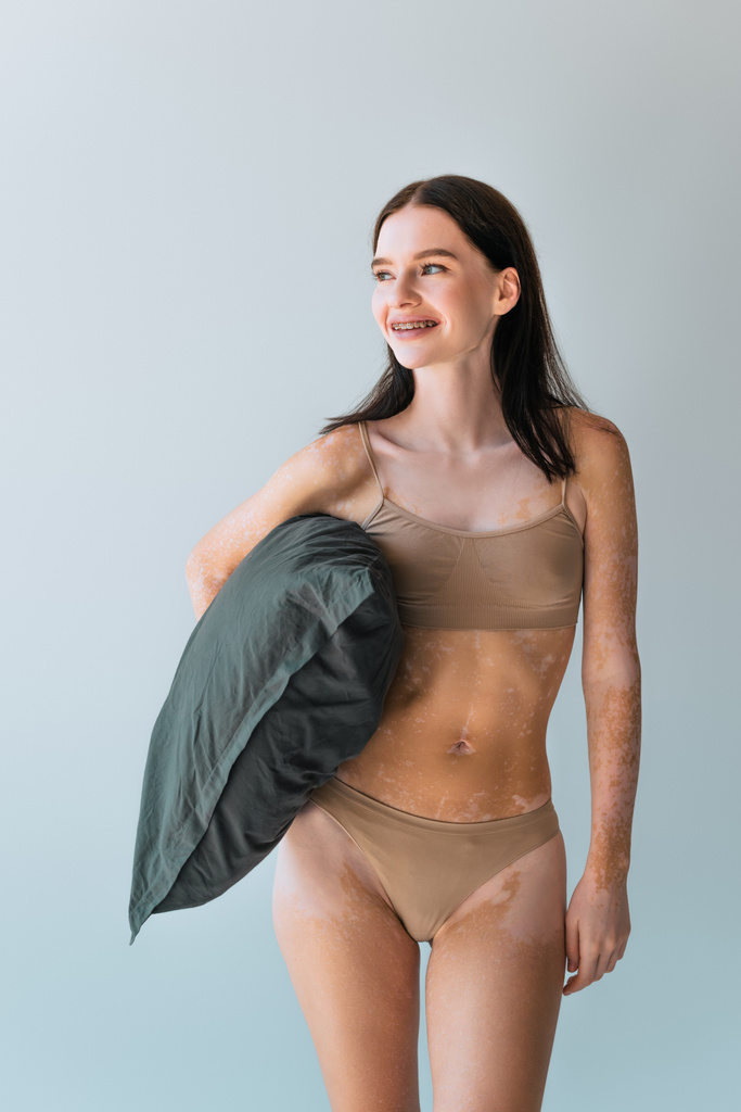 happy young woman with vitiligo and braces standing in beige lingerie and holding pillow isolated on grey - Photo, Image