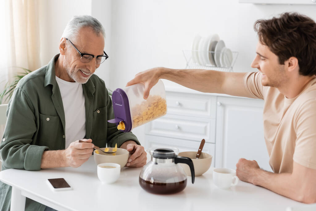 młody człowiek wlewając płatki kukurydziane do miski w pobliżu szczęśliwy tata i dzbanek do kawy w kuchni - Zdjęcie, obraz