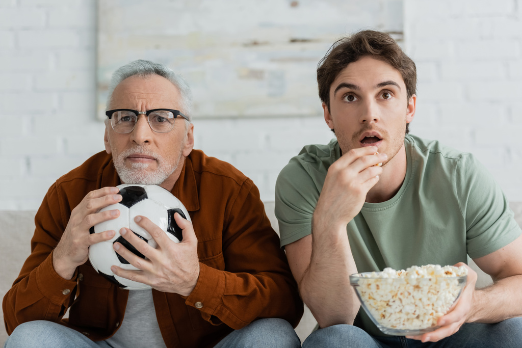 напряжённый взрослый мужчина держит футбольный мяч рядом с сыном, поедая попкорн во время просмотра чемпионата по телевизору - Фото, изображение