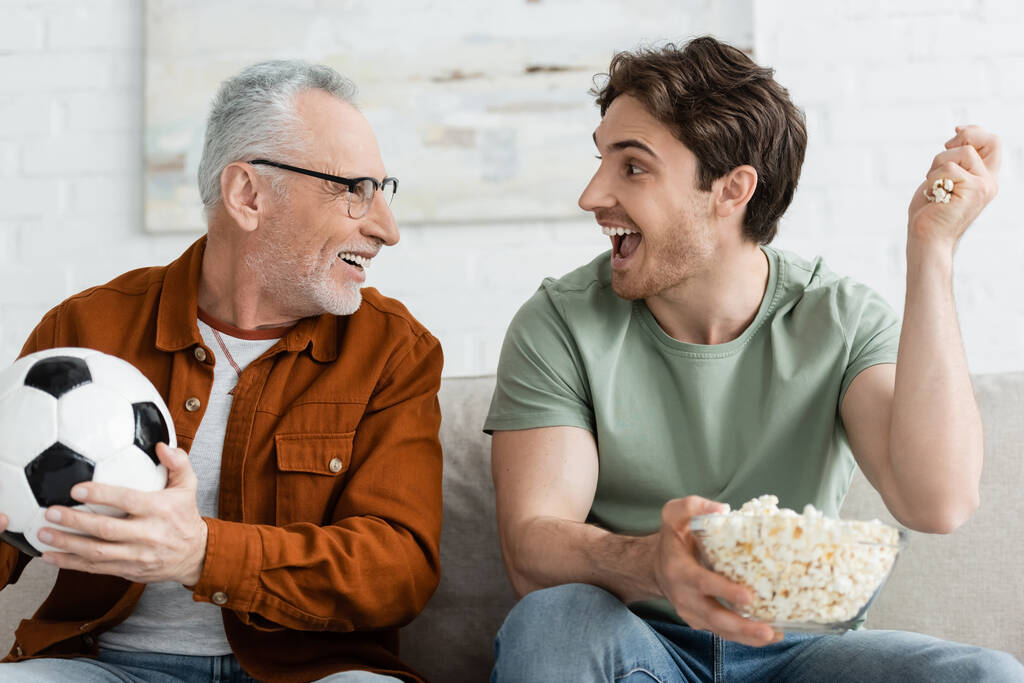 podekscytowany mężczyzna z popcornem i szczęśliwy ojciec z piłką nożną patrząc na siebie w salonie - Zdjęcie, obraz