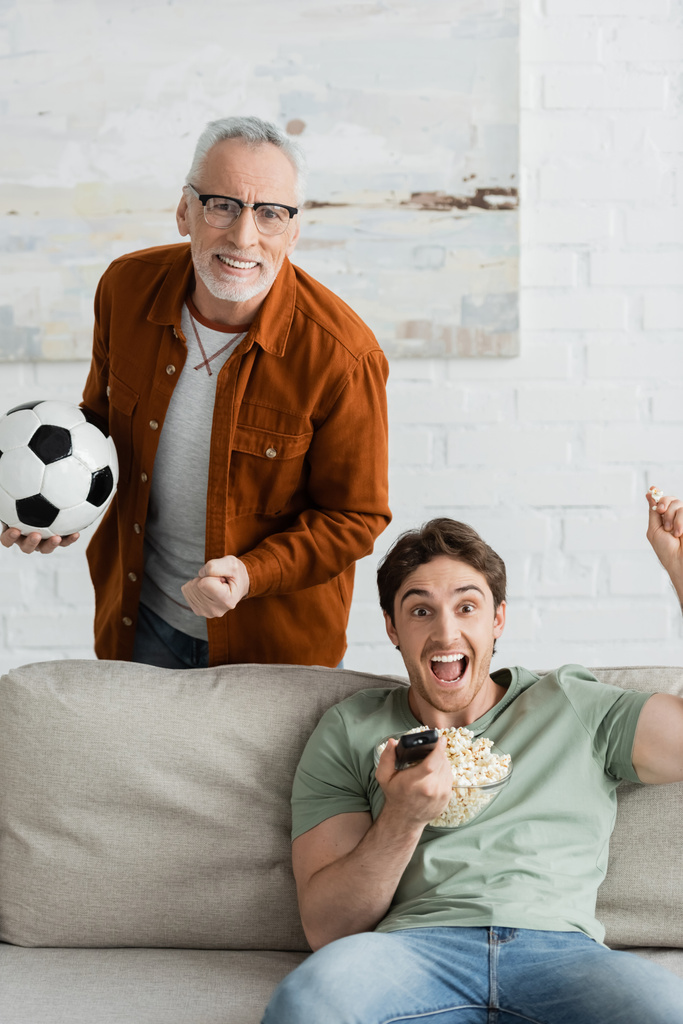 χαρούμενος γκριζομάλλης άντρας που κρατάει μπάλα ποδοσφαίρου και χαίρεται σχεδόν ενθουσιασμένος γιος που κρατάει τηλεχειριστήριο και ουρλιάζει στον καναπέ - Φωτογραφία, εικόνα
