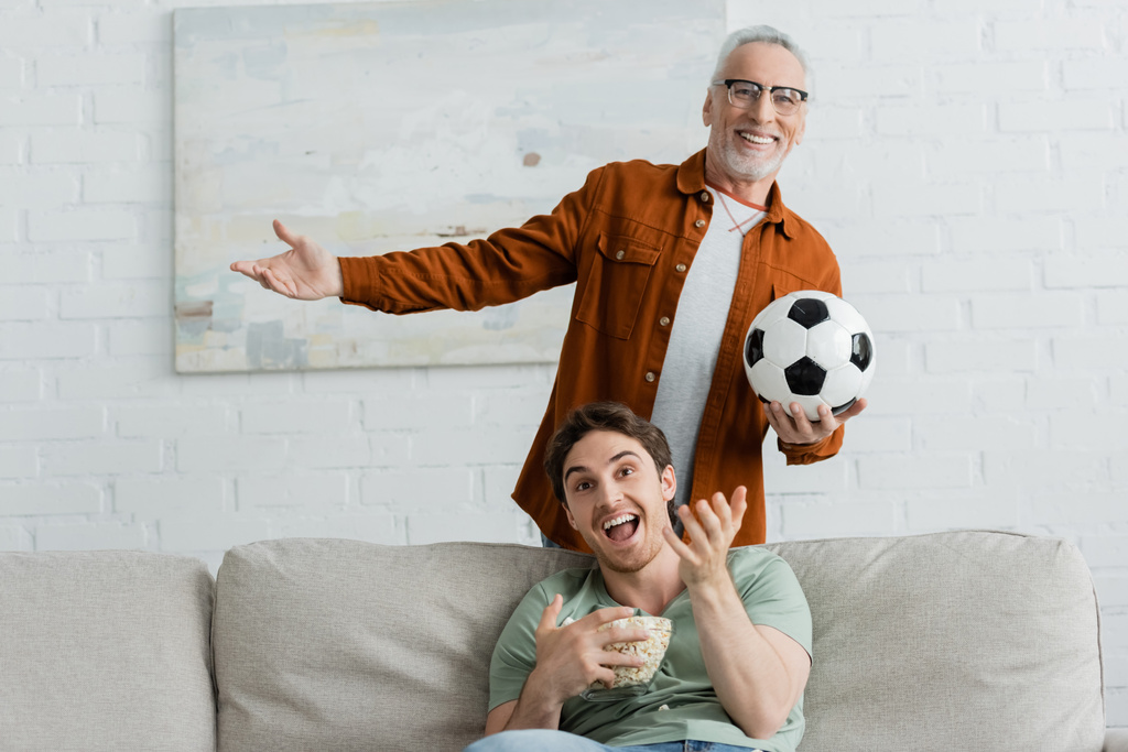 homme heureux avec ballon de football et fils gai avec bol de pop-corn geste tout en regardant match de football à la télévision - Photo, image