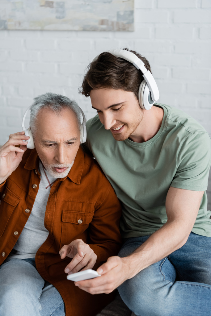 若い笑顔の息子の近くのスマートフォンを指している無線ヘッドフォンの灰色の髪の男 - 写真・画像