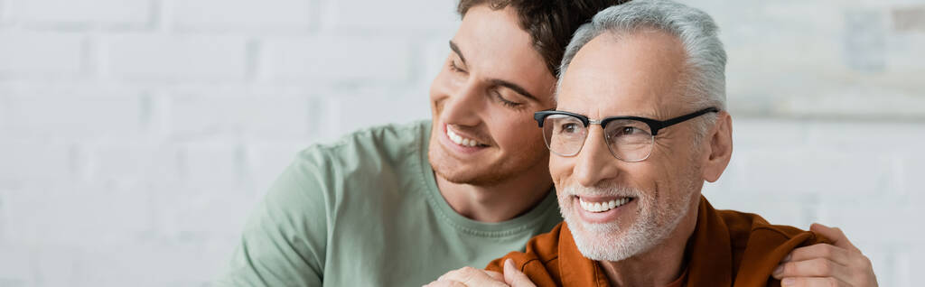 cheerful young man with closed eyes hugging bearded and mature father in eyeglasses, banner - Photo, Image