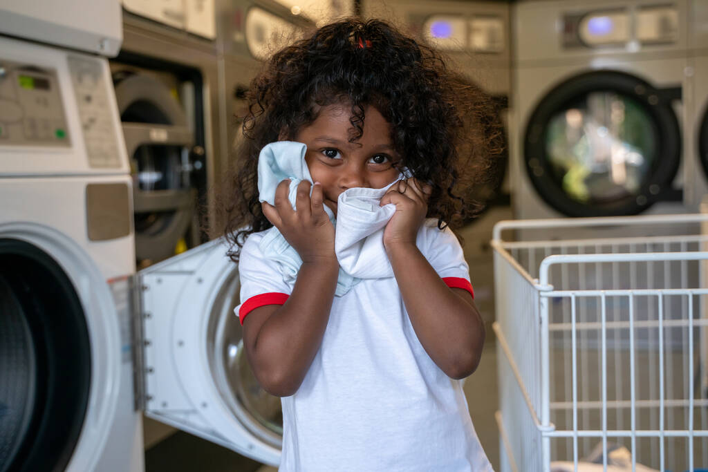 Lockiges kleines Mädchen, das allein neben einer Waschmaschine steht und ihr Gesicht mit Kleidung bedeckt - Foto, Bild