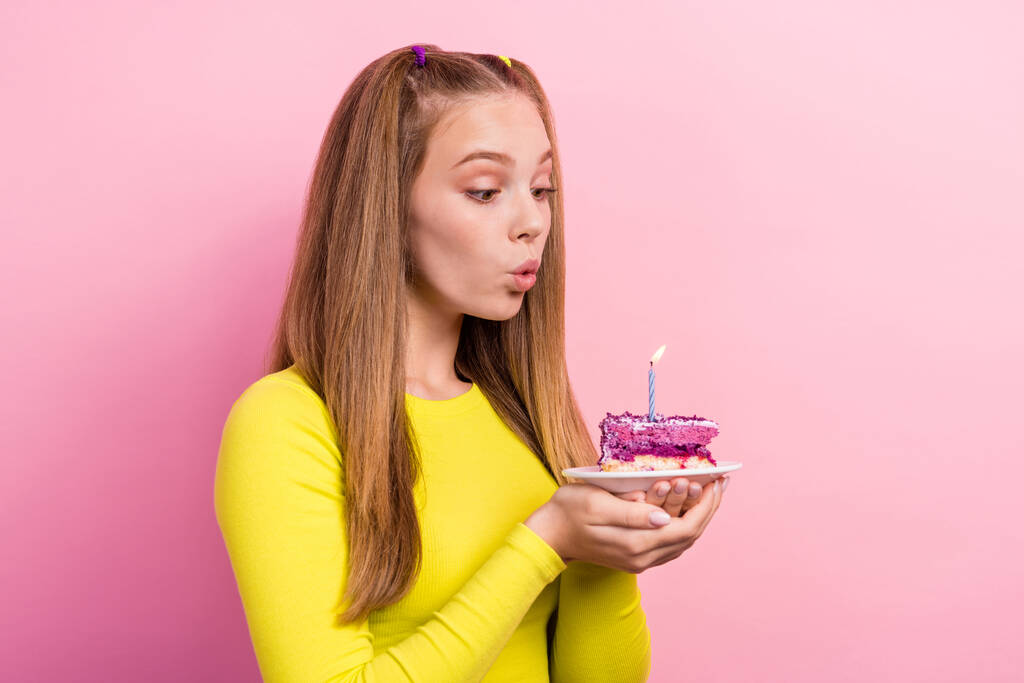 Fénykép fényes álmodozó iskolás lány viselni sárga felső gazdaság torta darab lemez, amelynek születésnapi kívánsága elszigetelt rózsaszín háttér. - Fotó, kép
