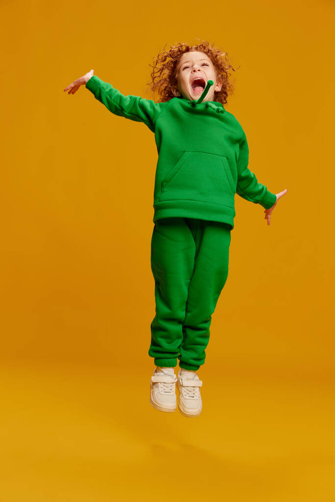 Portrait of cute little girl, child with curly red hair cheerfully jumping and laughing isolated on yellow background. Concept of childhood, emotions, lifestyle, fashion, happiness. Copy space for ad - Photo, Image