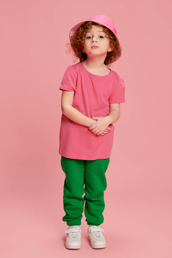 Portrait of cute stylish little girl, child with curly red hair posing in panama isolated over pink background. Concept of childhood, emotions, lifestyle, fashion, happiness. Copy space for ad - Photo, Image