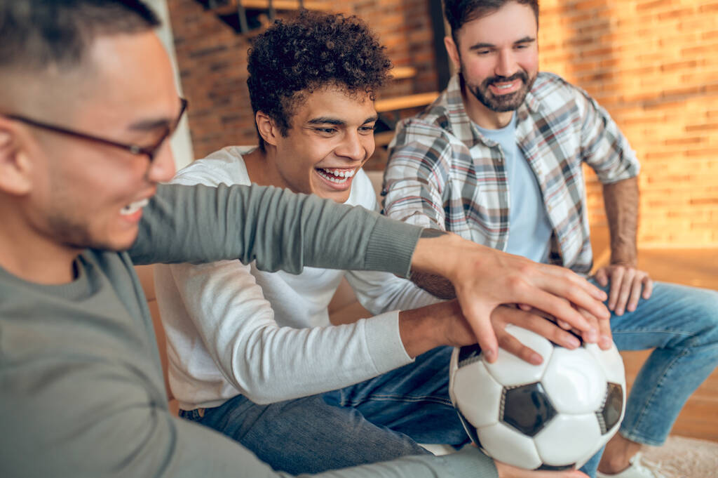 Sorridente allegro giovane uomo ei suoi amici soddisfatti mettendo le mani sul pallone da calcio - Foto, immagini