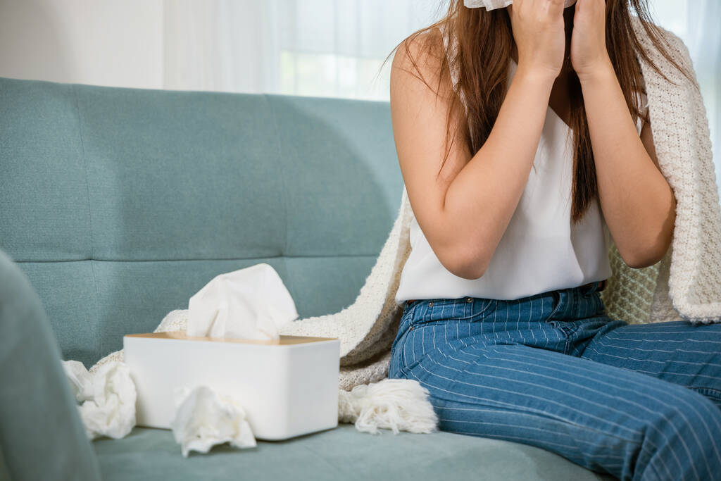 Mujer enferma sentada debajo de una manta en el sofá y estornuda con papel de seda en la sala de estar, fiebre atrapada en el frío, mujer joven asiática que está enferma sonándose la nariz estornudos en el tejido en casa, atención médica - Foto, imagen