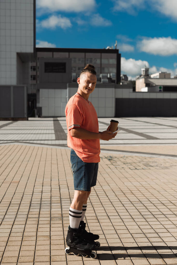 Mladý bruslař drží smartphone a kávu jít při pohledu na kameru venku  - Fotografie, Obrázek