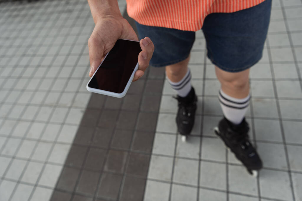 Обрезанный вид роликовой фигуристки, держащей смартфон с чистым экраном на открытом воздухе  - Фото, изображение